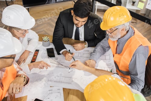 Preview: Multicultural colleagues in suits and helmets sitting at table and looking at blueprints.