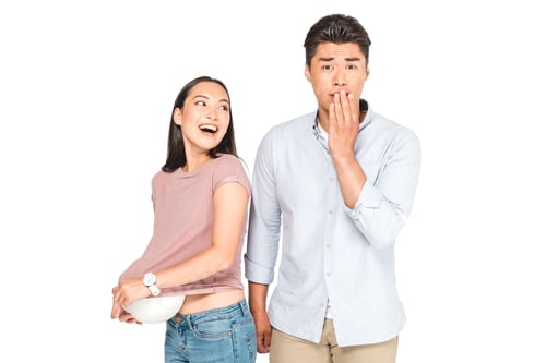 Preview: shocked asian man looking at camera while girlfriend getting ball out of t-shirt isolated on white