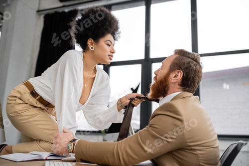 Preview: side view of passionate african american woman sitting on desk and pulling tie of bearded coworker