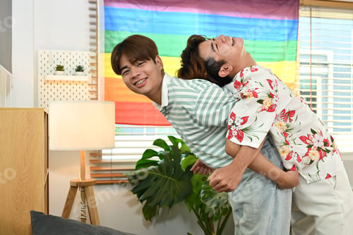 Preview: Laughing gay male couple giving piggyback ride for each other in living room with rainbow.