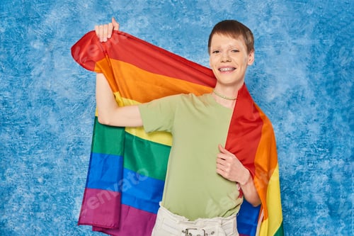 Preview: Cheerful gay man in casual clothes smiling and holding lgbt flag and looking at camera during pride