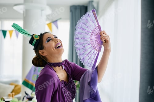 Preview: Young cheerful woman in Mardi Gras make-up and costume having fun at home.