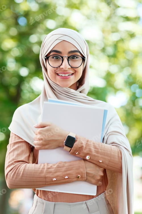 Preview: Cheerful glad cute young arab muslim female student in hijab and glasses holds books, ready for
