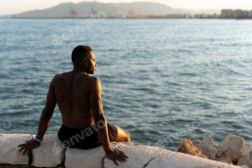 Preview: Sportsman sitting on the ocean coast and looking away while enjoying of the sunset