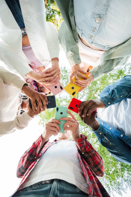 Preview: Vertical low angle view of group of friends standing in circle, all using cellphones outdoors