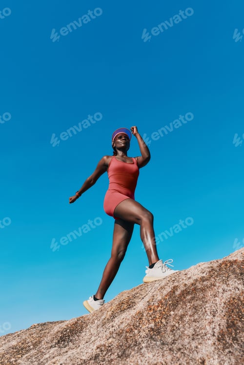 Preview: Young woman in sporty outfit standing on a rock against a clear blue sky