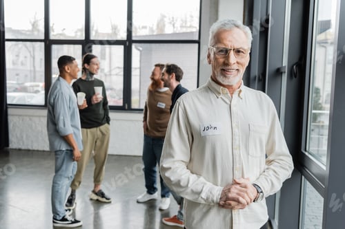 Preview: Smiling middle aged man looking at camera near blurred group during alcoholics meeting in rehab
