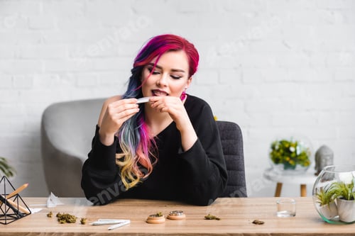 Preview: attractive girl licking joint with medical marijuana while sitting behind table in living room