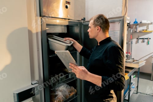 Preview: chef stands in kitchen in a black uniform and looks through what is on the list in the refrigerator