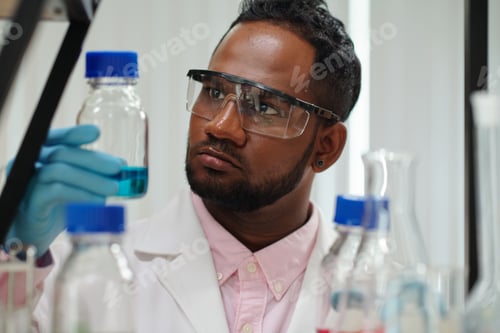 Preview: Serious young male clinician looking at glassware containing blue liquid