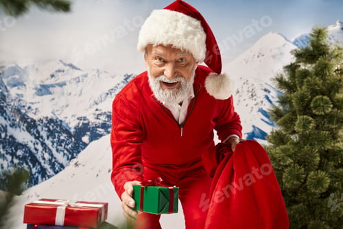 Preview: playful Santa surrounded by presents showing one at camera with slightly open mouth, winter concept