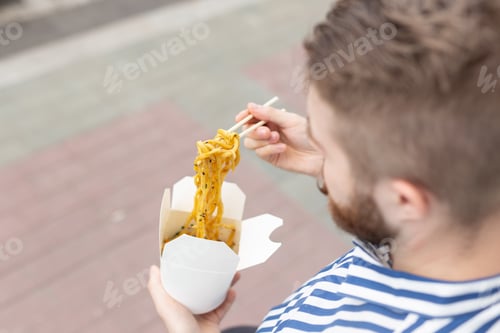 Preview: Young male hipster with a mustache and beard is eating chinese noodles with wooden chopsticks from a