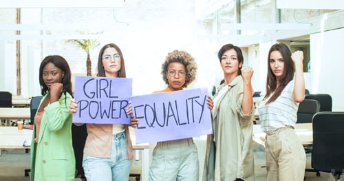 Preview: Group of five multiethnic women in an office looking at the camera hold up posters with the slogan: