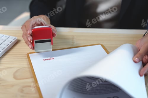Preview: Close-up of female hands stamping approved on a document with red ink at office desk, concept of suc