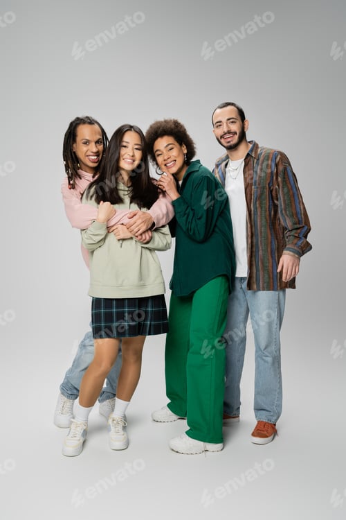 Preview: full length of joyful and stylish multicultural friends embracing and looking at camera on grey