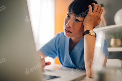 Preview: Asian female nurse in blue scrubs, looking puzzled while using a laptop.