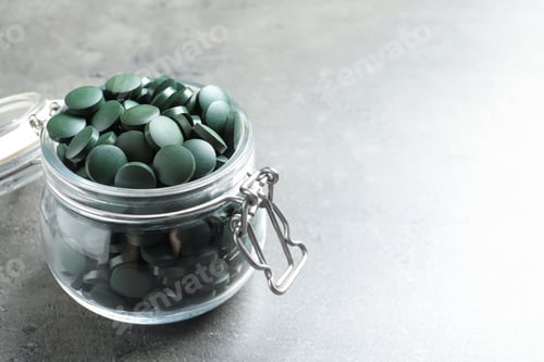 Preview: Glass jar with green spirulina pills on grey table, space for text