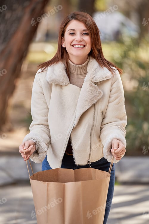 Preview: Woman looking at camera and smiling while holding a shopping bag outdoors.