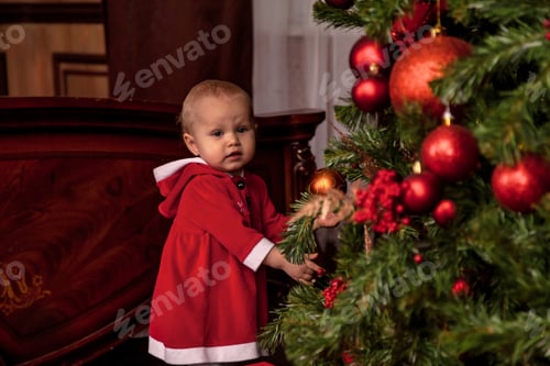 Preview: One-year-old girl disguised Santa Claus on background of Christmas tree decorated indoors