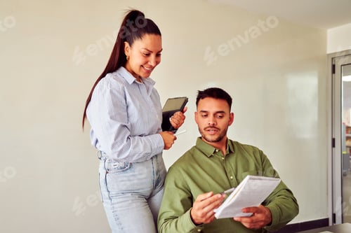 Preview: Businesswoman holding tablet helping businessman reading notes in office