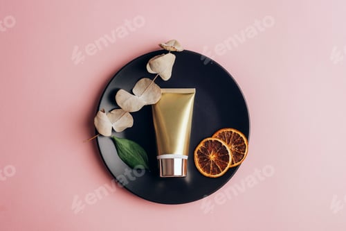 Preview: Golden cream tube and dry orange slices, eucalyptus leaves on black plate on a pink background.