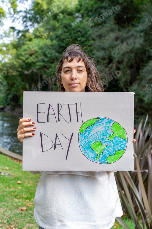 Preview: Vertical shot of a young woman holding a sign for Earth Day by the river