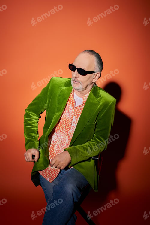 Preview: senior and confident bearded man sitting on chair and looking away on red and orange background