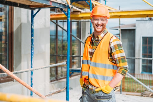 Preview: handsome smiling builder in reflective vest and hard hat looking at camera at construction site