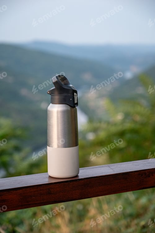 Preview: Stainless steel water bottle or thermos with black lid on wooden railing, blurred background