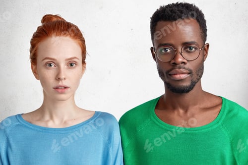 Preview: Serious dark skinned African male in spectacles stands next to his red haired beautiful Caucasian fe