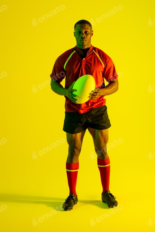 Preview: Portrait of african american male rugby player with rugby ball over yellow lighting
