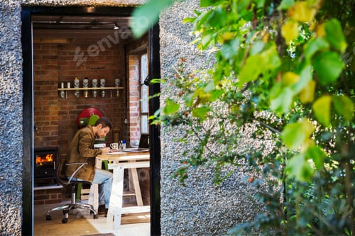 Preview: View through an open door, a designer seated in his leatherwork workshop at a desk, drawing.