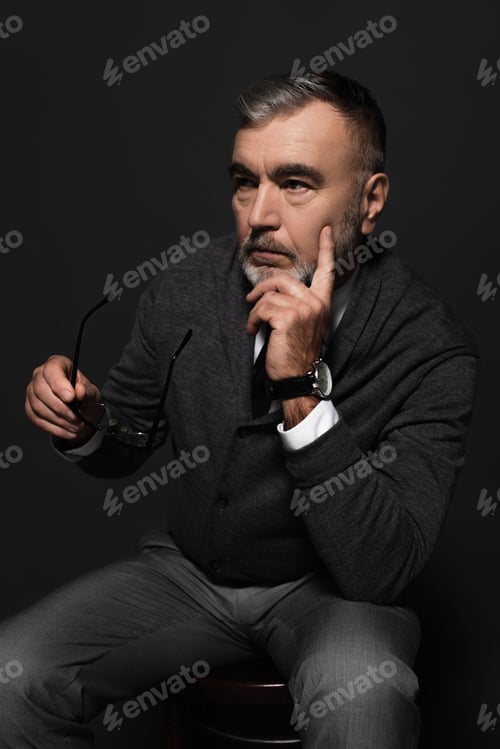 Preview: pensive man holding eyeglasses and looking away while thinking on dark grey