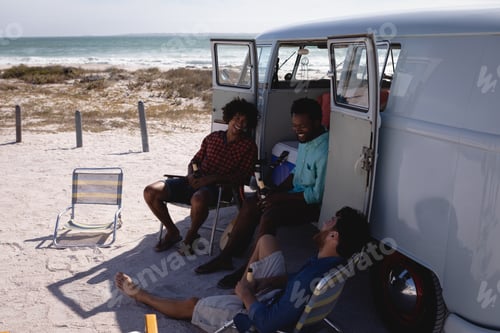 Preview: Front view of happy young multi ethnic men relaxing near camper van at beach in the sunshine
