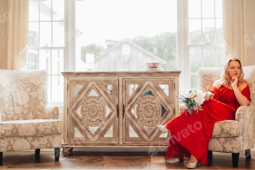 Preview: Portrait of a young millennial woman in red dress sitting in deep thought on a sofa in a venue