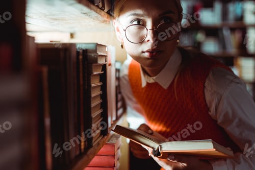 Preview: beautiful woman in glasses holding book and looking away in library