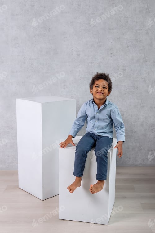 Preview: African American little boy in blue shirt, jeans sits on white large cubes, barefoot on a gray wall