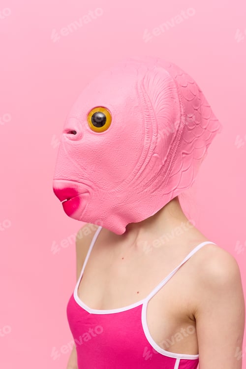 Vista previa: A woman in a pink fish head mask stands in profile against a pink background and looks into the