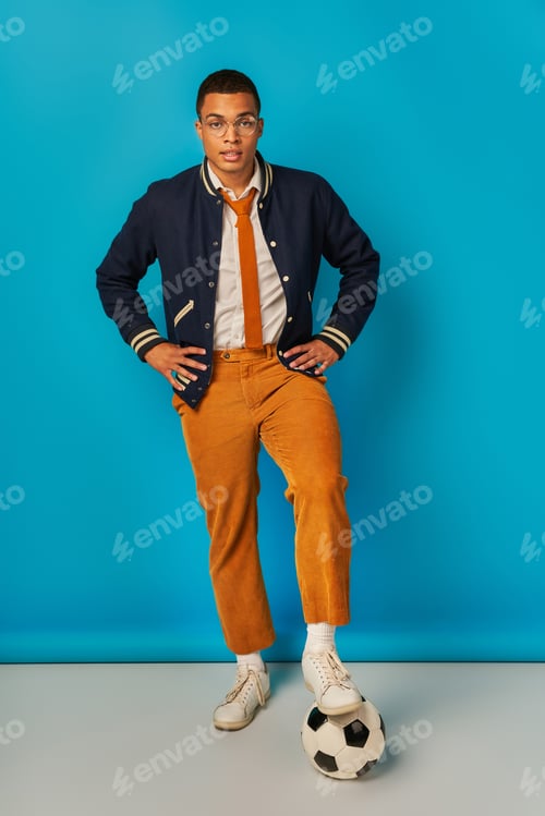 Preview: african american student in stylish clothes stepping on soccer ball and looking at camera on blue
