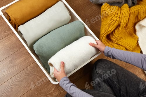 Preview: The hands of a woman neatly folding knitted clothes into a metal laundry basket