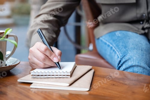 Preview: Closeup image of a woman's hand writing on blank notebook with coffee cup on wooden table