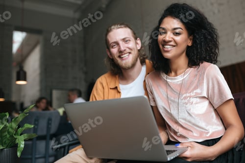 Preview: Young man and girl sitting at the restaurant and happily looking in camera with laptop in hands