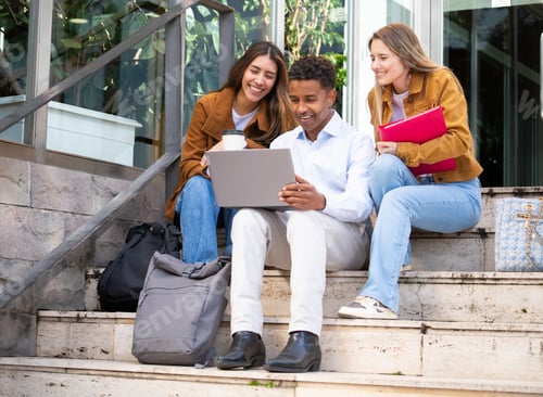 Preview: Happy diverse students using laptop and collaborating on project while sitting on stairs outdoors