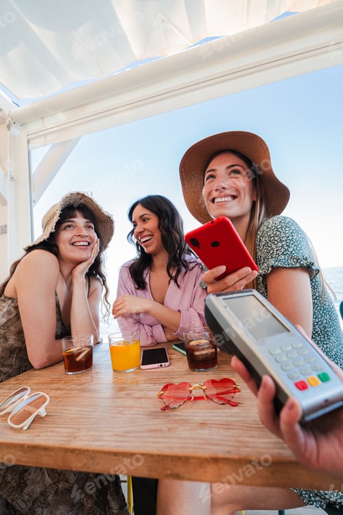 Preview: Vertical young group of only women paying the bill with a nfc cellphone in cocktail beach bar