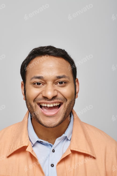 Preview: cheerful handsome indian man in orange jacket smiling straight at camera on gray background