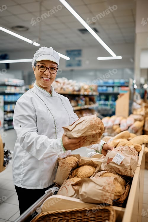 Preview: Happy baker arranging fresh bread in supermarket and looking at camera.