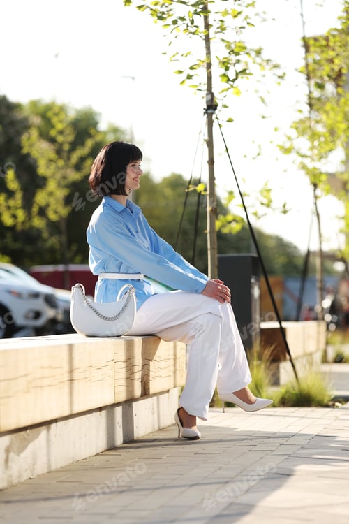 Preview: Elegant smiling brunette woman with short hair wears blue shirt, white pants and stylish bag sits on
