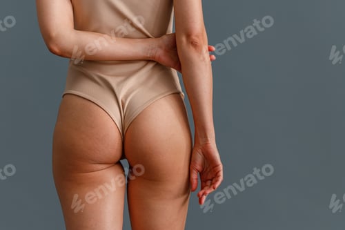 Preview: Back view of toned buttocks of woman in beige bodysuit standing isolated over gray studio background