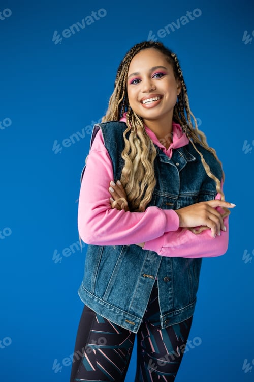 Preview: joyful african american woman in pink hoodie and denim vest on blue background, bold makeup