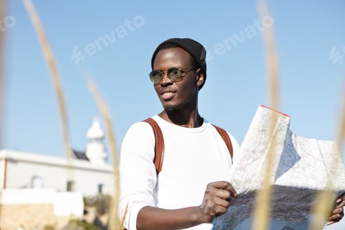 Preview: Summer portrait of handsome young black European man in trendy clothing using city guide while sight